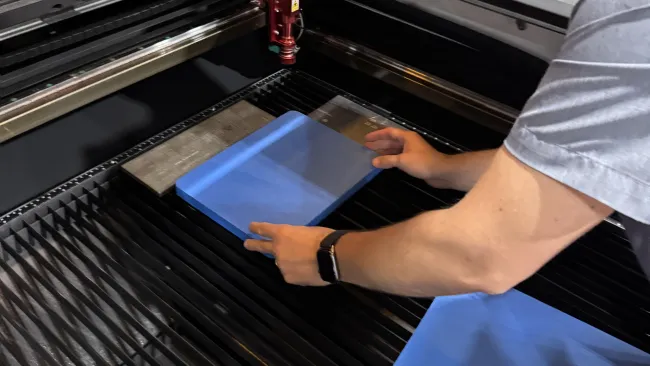 Person placing blue material inside laser cutting machine with red laser head and metal surfaces