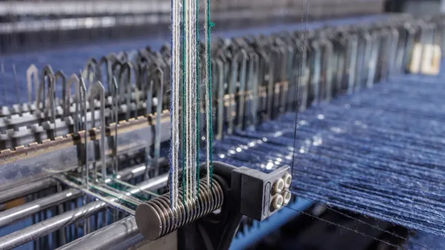 Close-up of industrial weaving machine with blue threads and metal components weaving fabric.