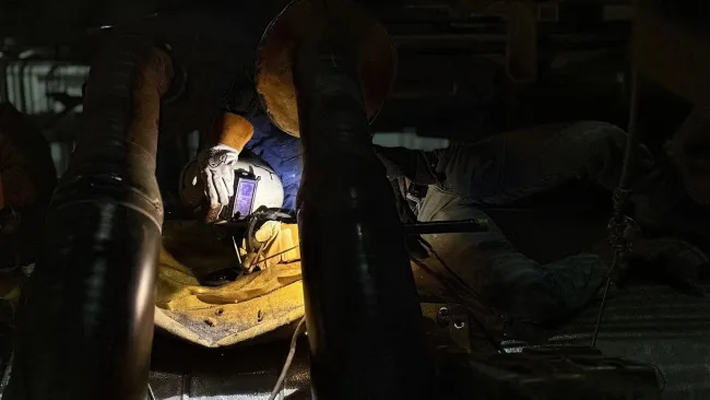 Welder in protective gear working under vehicle with bright torch light illuminating metal surfaces in dark environment