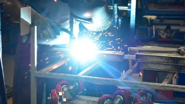 Worker wearing protective gear welding metal parts producing bright sparks in industrial workshop.