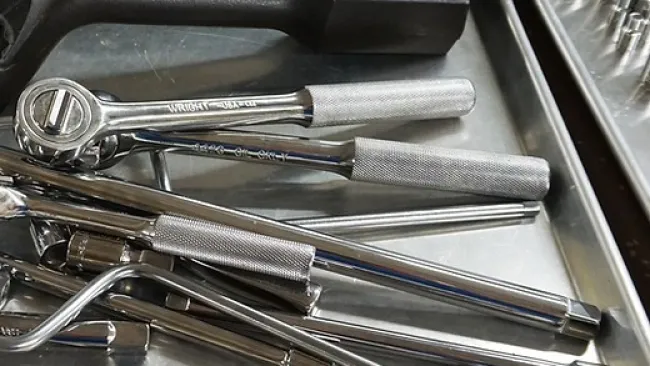 Close-up of various metal hand tools including ratchets, wrenches, and a black hammer on a steel tray.