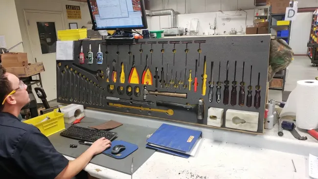 Man working at a workshop desk with organized tools hanging on a pegboard and a computer monitor above.