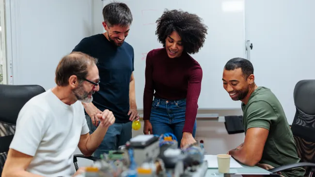 Four diverse coworkers collaborate over robotics project in modern office with whiteboard in background.