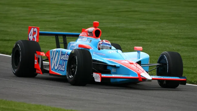Blue and red open-wheel race car number 43 speeding on a paved racetrack with grass in the background.