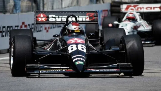 Black Formula 1 race car number 86 speeding on race track with another car behind during a race event.