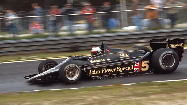 Black John Player Special Lotus Formula 1 race car speeding on track with driver in helmet and blurred crowd background.