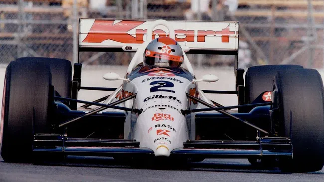Front view of a vintage IndyCar racing car with Kmart sponsorship and driver in orange helmet on race track.