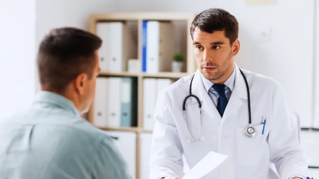 Doctor in white coat with stethoscope consulting male patient in modern medical office setting.