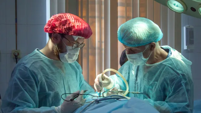 Two surgeons in surgical gowns and masks perform an operation under bright operating room lights.