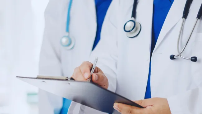 Two doctors in white coats with stethoscopes reviewing medical notes on a clipboard in a clinical setting