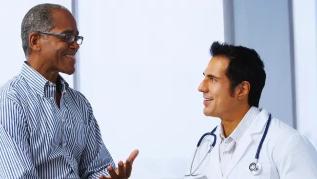 Doctor with stethoscope consulting a smiling male patient in a medical office, holding a tablet device.