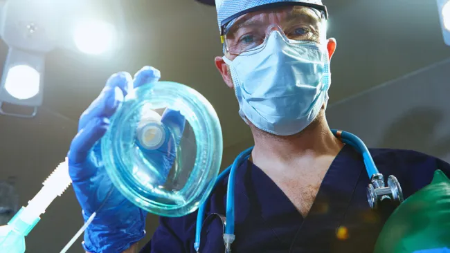 Surgeon in mask and gloves holding an oxygen mask preparing for medical procedure in operating room.