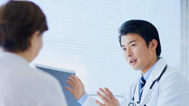 Doctor in white coat explains medical information to patient in bright consult room with computer.