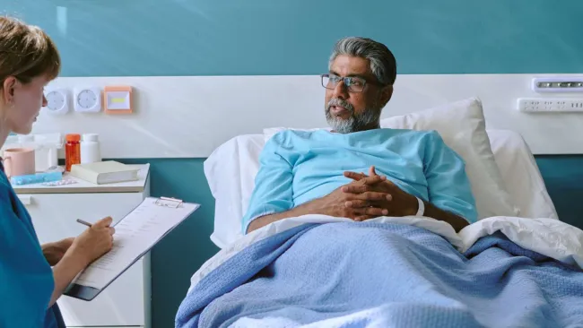 Male patient lying in hospital bed talking to nurse taking notes on clipboard in medical room