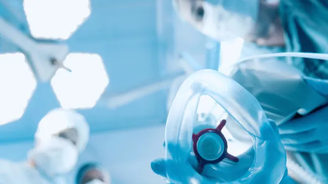 Medical team wearing masks in operating room holding anesthesia mask toward camera under bright surgical lights