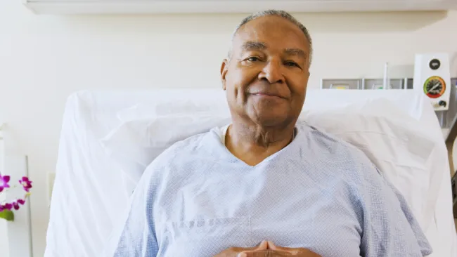 Smiling elderly man resting in hospital bed wearing light blue patient gown with folded hands.