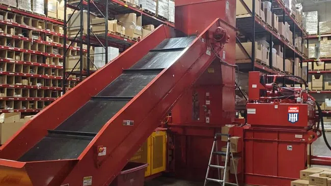 Red industrial conveyor belt system in a warehouse surrounded by shelves stacked with boxes and packages.