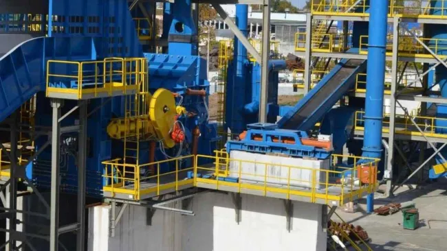 Industrial facility with blue and yellow machinery, platforms, and conveyor systems on a sunny day.