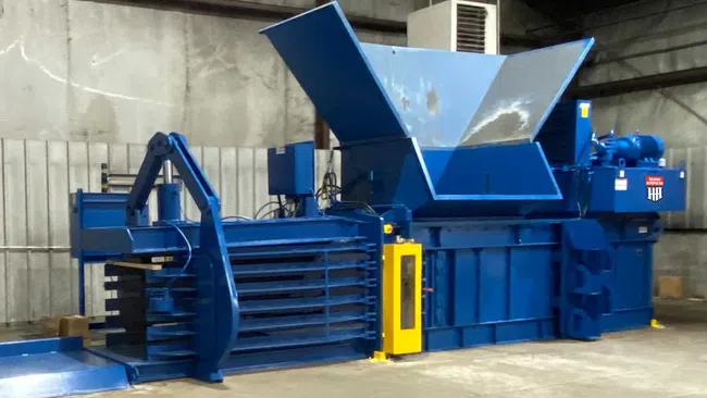 Large blue industrial baler machine with open hopper inside a warehouse setting.