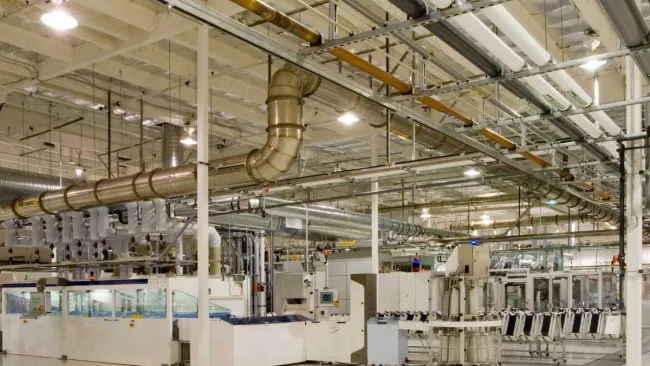 Spacious clean factory floor with industrial machinery and extensive overhead ventilation piping system.