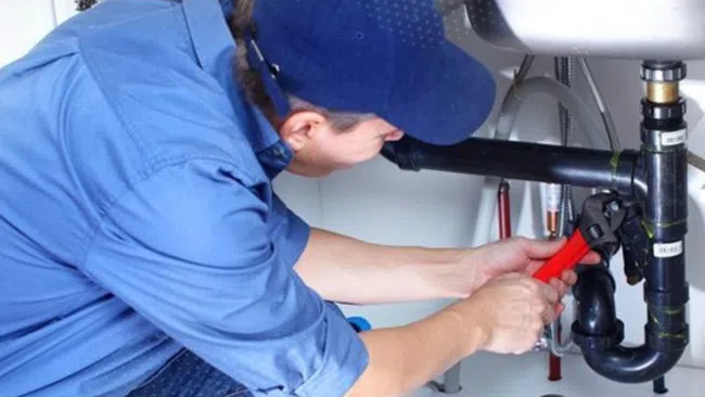 Plumber in blue uniform fixing black pipes under a sink using a red pipe wrench in a white room.