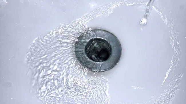 Tall gray water heater installed in a small utility closet with exposed pipes and concrete floor.