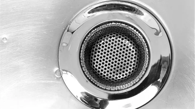 Close-up of a shiny metal sink drain with perforated strainer and reflective surface under light.