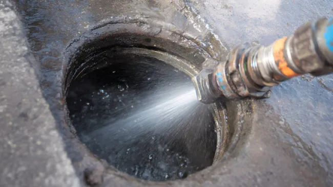 High-pressure water jet cleaning the inside of a concrete drain or sewer pipe opening outdoors.