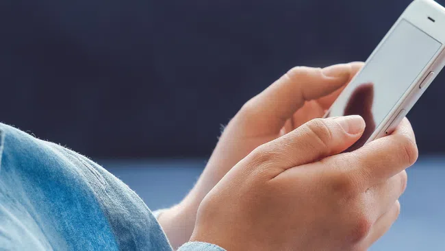 Person wearing denim jacket using a white smartphone with blurred blue background.