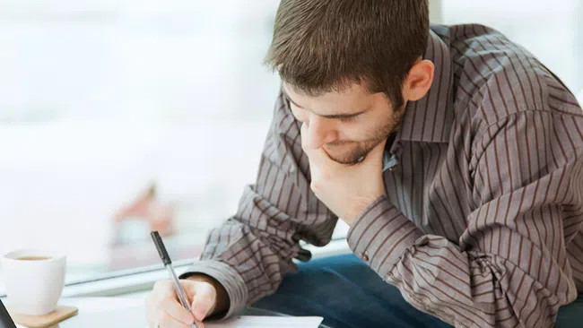 Man in striped shirt writing on paper at glass table with laptop and coffee cup by window