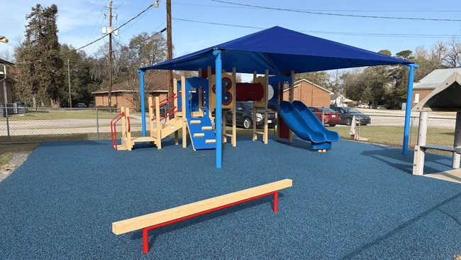 Children's playground with blue rubber flooring, slides, climbing structures, and a blue shade canopy under clear sky.