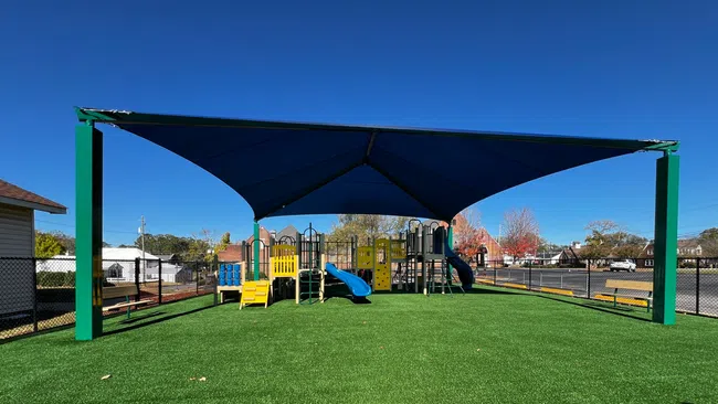 Children's playground with slides and climbing structures under a large blue shade canopy on artificial grass.