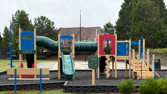 Colorful playground with slides, climbing structures, and benches in a park setting with trees and houses behind.