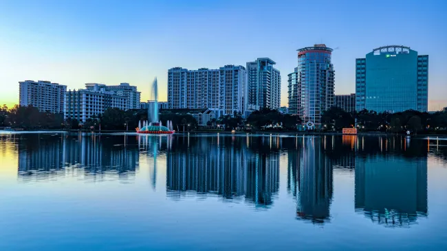 City skyline with modern buildings and a fountain reflected on calm lake water at sunset.