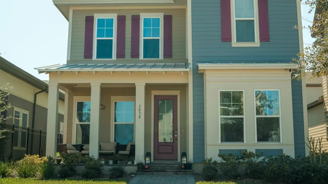 Two-story suburban house with blue siding, purple shutters, front porch, and well-kept lawn on sunny day.