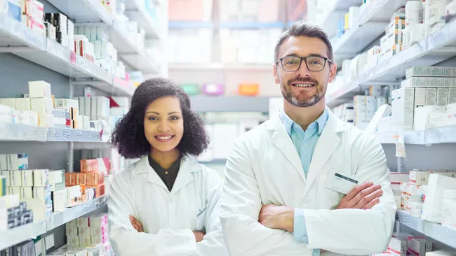 Two pharmacists in white coats standing confidently in a pharmacy aisle with shelves full of medication boxes.