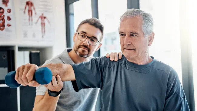 Senior man lifting blue dumbbells with assistance from trainer during a rehabilitation exercise session.