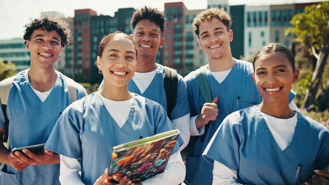 Group of diverse nursing students smiling outside in scrubs holding books and tablets with city buildings in background