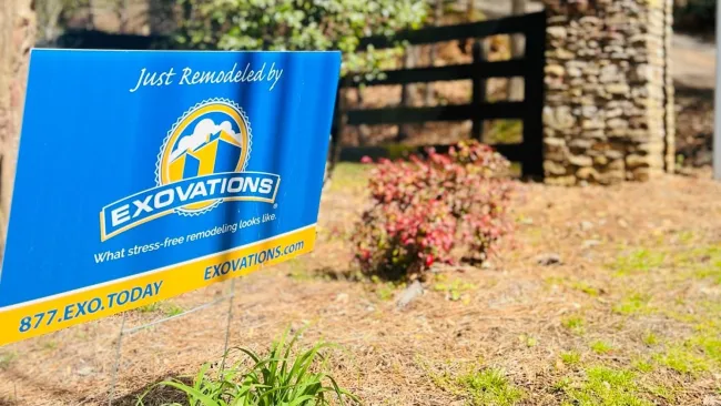 Blue and yellow yard sign for Exovations remodeling company in a garden with plants and a stone pillar fence