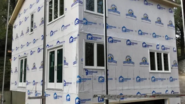 Two-story house under construction with windows installed and wrapped in white weatherproof barrier material.