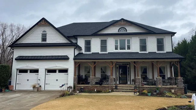 Two-story white house with black roof, large front porch, two-car garage, and a dry lawn under cloudy sky
