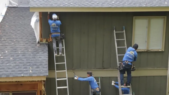Four construction workers in blue shirts install siding on a house using ladders and tools outdoors.