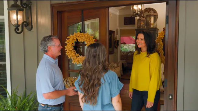 Smiling woman in yellow welcomes two visitors at a wooden front door decorated with a yellow wreath