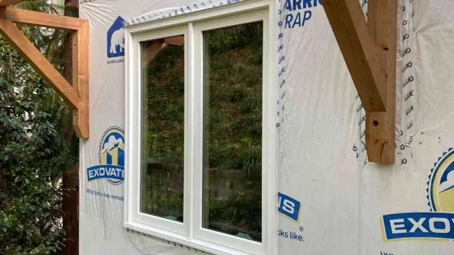 White double-pane window installed on a house under construction with wood framing and bushes below.