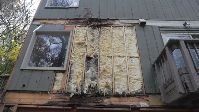 Damaged exterior wall shows exposed insulation and wood rotting beneath siding near window and balcony.