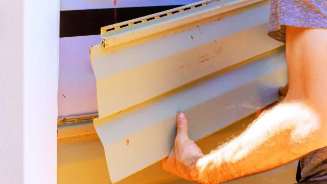Person installing beige vinyl siding on exterior wall with a tool belt visible at the waist.