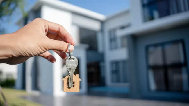 A hand holding a house-shaped key in front of a modern house.
