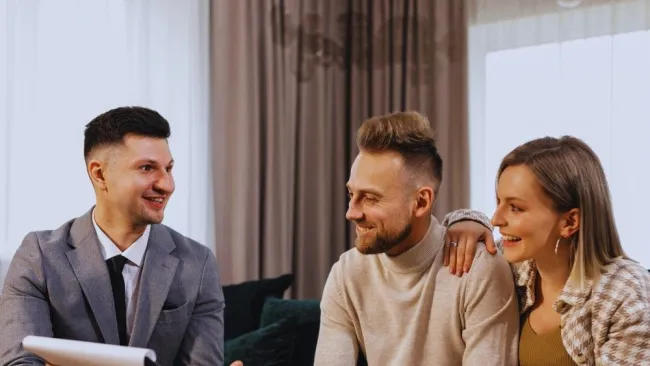 Couple smiling and discussing with a male consultant holding documents in a cozy living room setting.