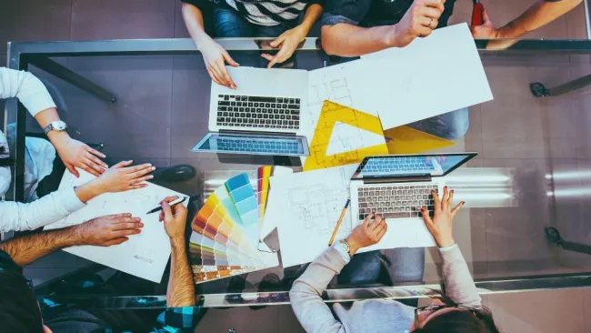 Overhead view of a team collaborating with laptops, color swatches, and architectural plans on a glass table