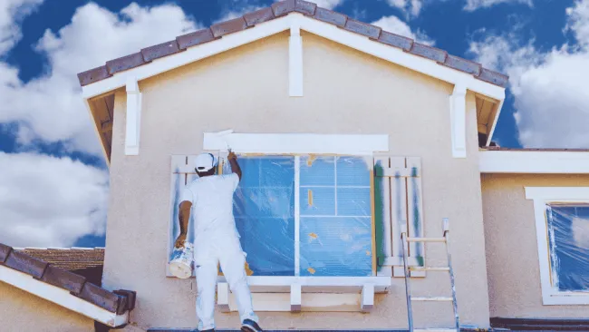 Painter in white outfit painting the exterior of a house under blue cloudy sky with a ladder propped nearby